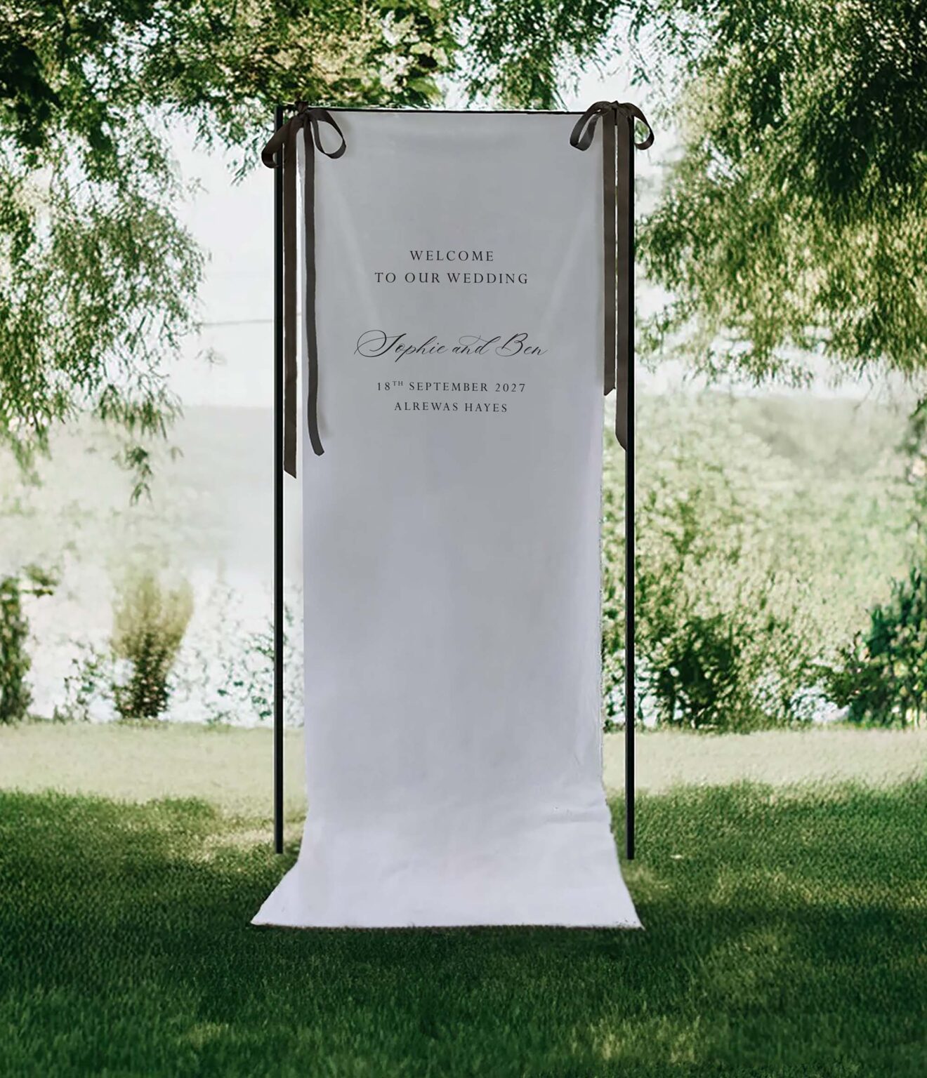 a linen welcome to our wedding sign with black satin ribbon. the stand it is on is black and it stands in a field overlooking a lake
